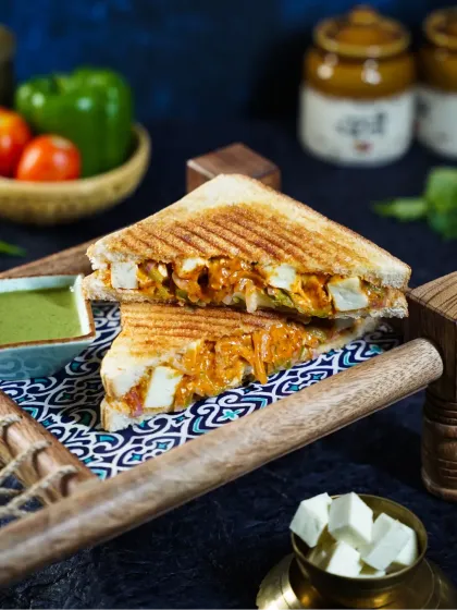 A grilled paneer tikka sandwich, cut in half to show the generous, creamy filling. The dark, moody styling with fresh ingredients in the background makes it look incredibly tempting.