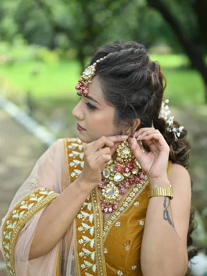 A close-up of a model adjusting her earring, wearing a mustard yellow lehenga and a heavy Kundan necklace with pink and green stones.