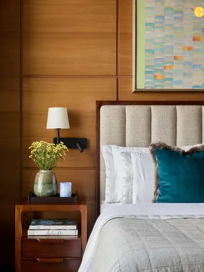 The master suite in 'Pad by the Bay' combines warm wood paneling with a comfortable, upholstered headboard. A pop of color from the teal velvet cushion and the abstract painting above the bed adds a touch of personality to the serene space.