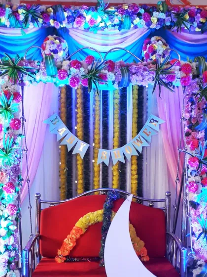 A close-up of a decorated swing for a baby shower. The frame is wrapped in colorful flowers and lights, with a "Baby Shower" banner hanging in the background.