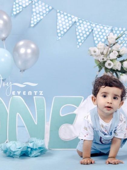 This is a classic blue-themed cake smash photoshoot I arranged for a baby boy. The setup includes a light blue backdrop, a large 'ONE' sign, silver and blue balloons, and a polka-dot bunting banner.