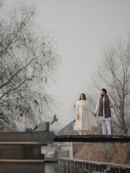 A serene portrait of a couple standing on a wooden bridge over the calm waters of Dal Lake. The misty, ethereal atmosphere of Kashmir provides a perfect setting for romantic and soulful pre-wedding photography.