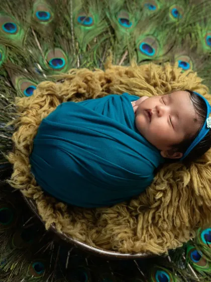 A close-up on the peacefully sleeping baby, showing the soft texture of the wrap and the delicate headband against the backdrop of peacock feathers.