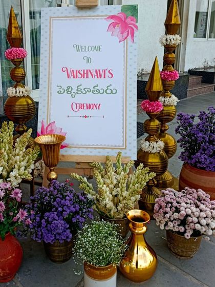 A welcome sign for a Pellikuthuru ceremony, adorned with traditional brass elements and beautiful purple and white flowers. We celebrate every ritual with style.