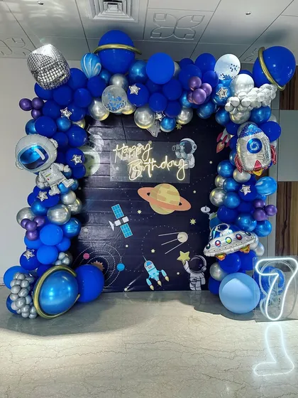 An out-of-this-world space theme birthday party for a 7-year-old. This detailed setup includes a galaxy backdrop, a full balloon arch in shades of blue and silver, and fun foil balloons shaped like astronauts, rockets, and planets.