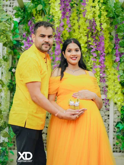 A happy couple holding tiny baby shoes, posing in front of a beautiful wall of flowers.