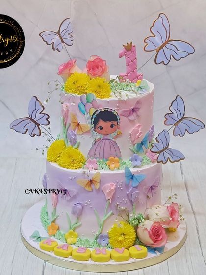 This two-tier fairy theme cake is a garden of delights, decorated with real flowers, paper butterflies, and a sweet edible baby picture for a personalized and magical touch.