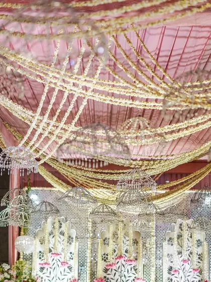 Looking up at the elaborate ceiling decor, with its crisscrossing garlands of mogra, draped fabrics, and hanging crystal elements. The design creates a sense of grandeur and celebration.