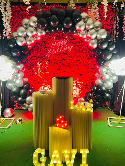 A bold birthday decoration with a red floral backdrop and a black and silver balloon arch. The bright lighting and marquee letters for 'GAVI' make the whole setup pop.