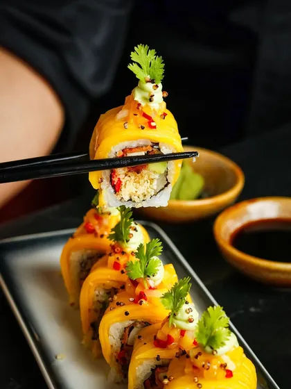 Chopsticks lifting a piece of mango-topped sushi, shot with dramatic, dark lighting to create a moody and sophisticated feel.