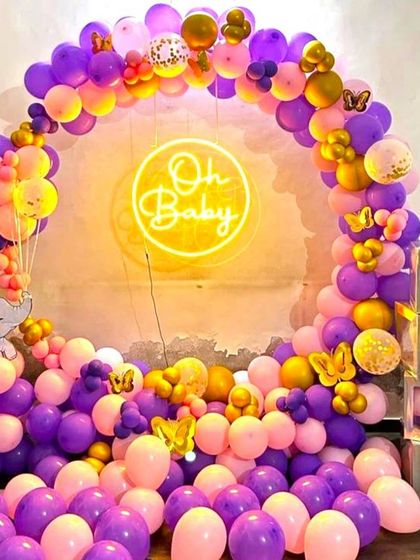 A vibrant baby shower decoration with a purple, pink, and gold balloon arch. The setup includes an 'Oh Baby' neon sign and transparent 'BABY' blocks, creating a beautiful photo corner.