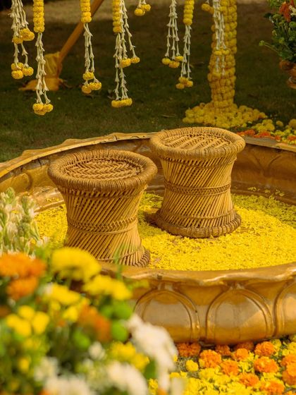 Traditional cane 'mooda' stools placed inside a large brass urli filled with flower petals. This detail combines rustic charm with classic elegance for a unique haldi setup.
