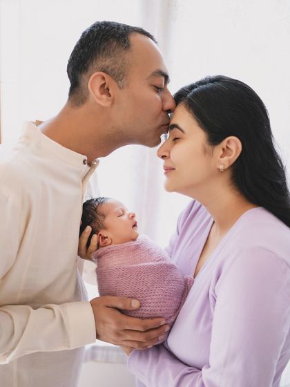 A tender family moment, with a father's loving kiss on his partner's forehead as they both hold their new baby.