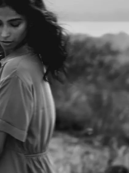 A soft, melancholic black and white portrait. The model is turned away, looking down, with the focus on the gentle light on her back and the soft, out-of-focus natural background.