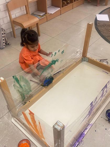 An overturned table wrapped in plastic becomes a three-dimensional canvas. This innovative setup invites the child to paint from different angles, developing spatial awareness and creative problem-solving skills.