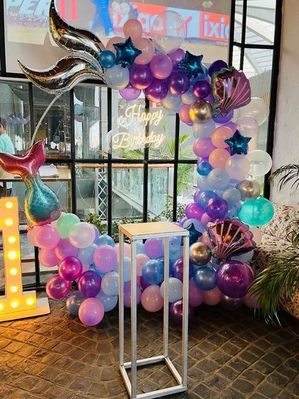 A magical mermaid-themed birthday party. The balloon arch in shades of purple and blue is adorned with large mermaid tail and seashell foil balloons.