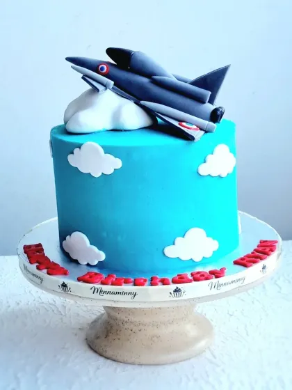 A fighter jet cake with a sky-blue buttercream base and fondant clouds. The jet itself is sculpted from gumpaste for a clean, detailed finish.