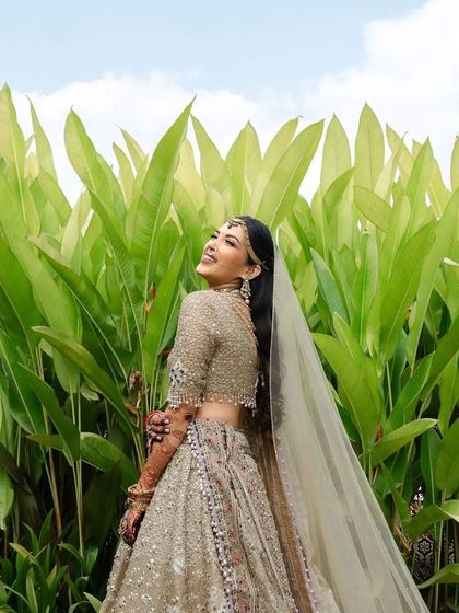 A happy bride against a lush green background, her stained henna visible on her arms.