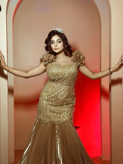 A full-length view of the golden sequined mermaid gown with a pleated trail. The floral details on the shoulders add a unique touch.