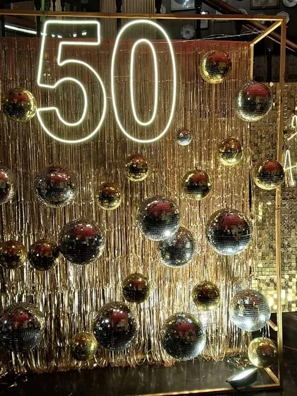 This wide shot shows the full scale of the 50th-anniversary setup. We combined a framed disco ball installation with a gold sequin wall and a 'Happy Anniversary' neon sign for maximum glamour and impact.