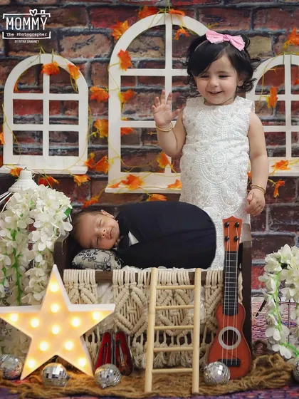 A proud big sister waves hello next to her sleeping newborn brother. This setup with the star light and brick background creates a warm and cozy atmosphere for a sibling photoshoot.