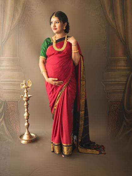 A graceful standing pose of a mom-to-be in a red saree, showcasing the baby bump beautifully in a traditional setting.