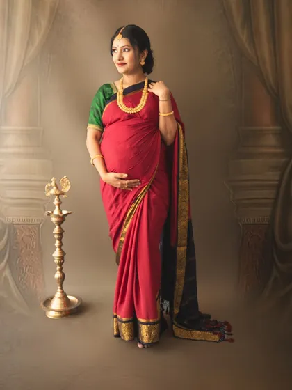 A graceful standing pose of a mom-to-be in a red saree, showcasing the baby bump beautifully in a traditional setting.