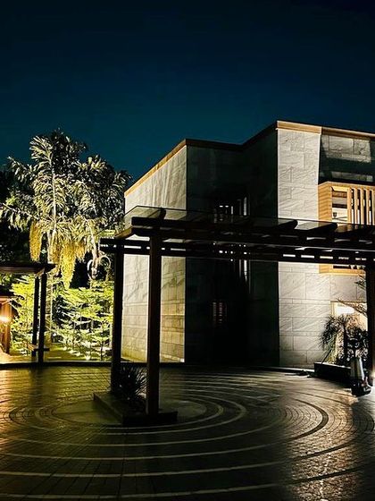 A night view of the farmhouse entrance, where the warm interior light contrasts with the cool tones of the exterior stone. The wooden pergolas add texture and depth to the facade.