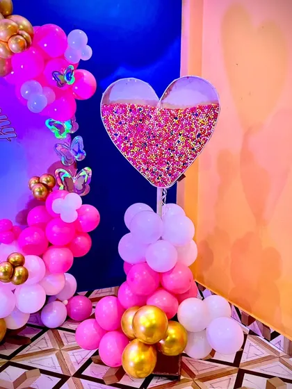A lovely decoration for a naming ceremony or baby shower. This setup includes a pink, white, and gold balloon garland and a unique heart-shaped stand filled with colorful sprinkles.