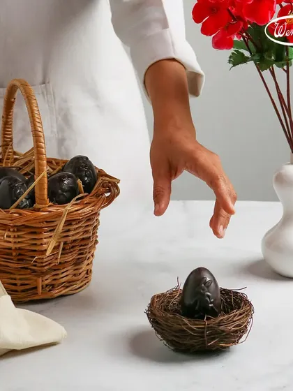 Getting ready for the Easter egg hunt. A hand reaches for one of our solid dark chocolate eggs, resting in a tiny nest, with a basket full of more eggs in the background.