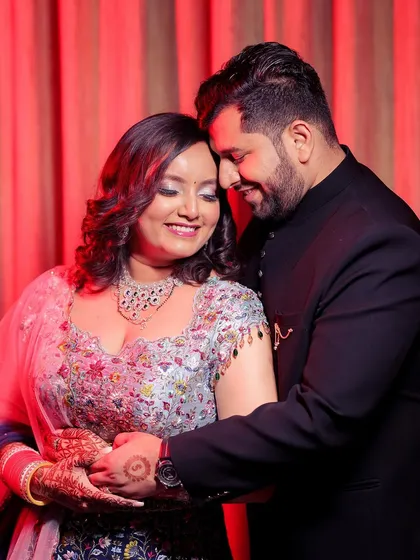 This is a warm, romantic portrait from an engagement celebration. The couple holds each other in a gentle embrace, their smiles showing their comfort and love. The red curtain backdrop adds a touch of drama and richness to the scene.