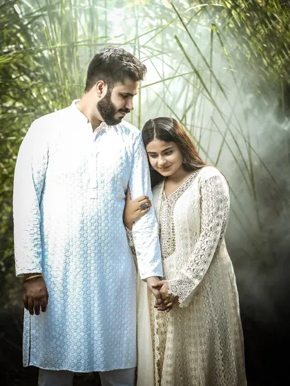 A quiet, intimate moment during a pre-wedding shoot. The couple is dressed in traditional white outfits, creating a soft and romantic mood amidst a natural, green setting.