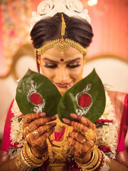 A close-up of a bride holding two hand-painted paan leaves before the Shubho Dristhi. The delicate white patterns on the green leaves create a stunning visual.