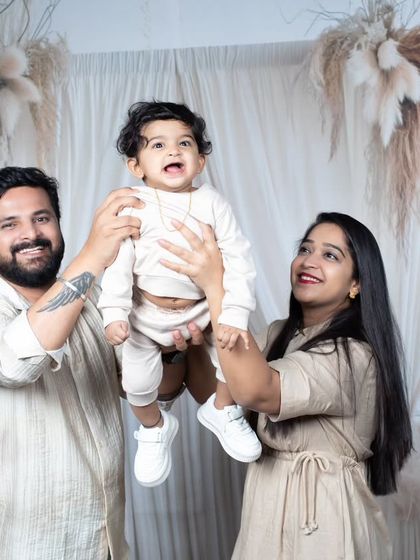 A joyful family of three, with parents lifting their baby boy in the air against a neutral-toned boho backdrop.