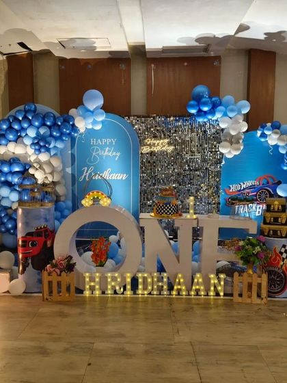 A high-speed Hot Wheels theme for a first birthday. The blue-themed setup includes branded backdrops, 'ONE' marquee letters, and plinths decorated with race car graphics.