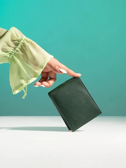 A hand delicately balancing a green leather wallet against a teal background. The composition is minimalist and elegant, and the color combination is fresh and modern.