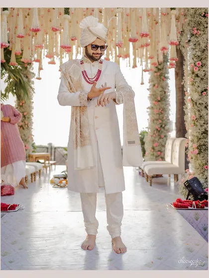 The groom awaits his bride, standing on a mirrored aisle that reflects the stunning floral decor. This setup highlights the grandeur of the entrance and the mandap.