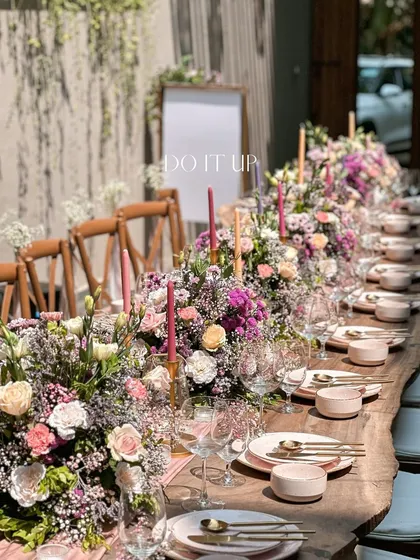 A long, rustic wooden table set for a bridal shower. The tablescape features a continuous runner of lush purple and pink florals, accented with taper candles, creating a vibrant and communal dining experience.