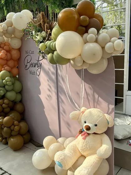 A cuddly teddy bear sits on a cloud of white balloons, holding a smaller balloon bunch. This floating effect adds a touch of whimsy to the earthy-toned setup.
