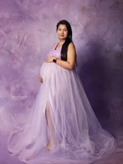 A full-length shot showcasing the beautiful drape of the lavender tulle maternity gown. This classic pose highlights the baby bump and the elegance of the expecting mother.