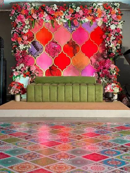 This vibrant Mehendi stage features a colorful Moroccan tile backdrop, framed by a lush arch of pink and orange flowers, and a modern olive green sofa.