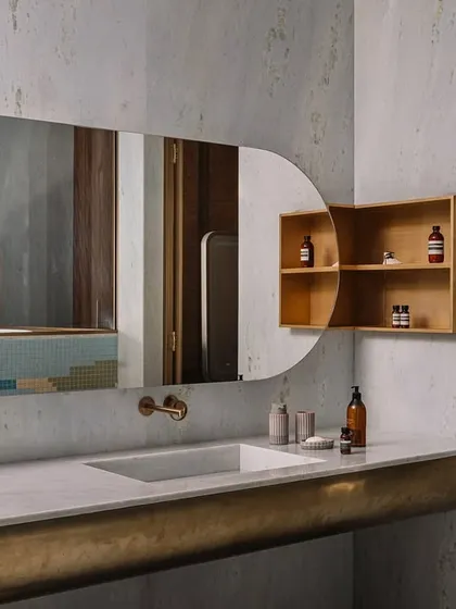 This bathroom vanity combines a sleek, curved brass base with a solid countertop and an integrated sink. Paired with a custom-shaped mirror and a simple wooden medicine cabinet, the design is a study in material contrast and clean, modern forms.