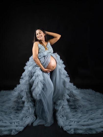 This dusty blue ruffled gown creates a spectacular circular train, perfect for a dramatic full-body portrait. The open-front design beautifully showcases the baby bump against a classic dark backdrop.
