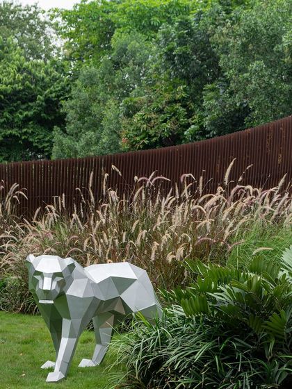 A geometric, silver-colored bear sculpture stands in a grassy area, with a curving Corten steel screen in the background. This juxtaposition of art and landscape creates a playful yet sophisticated focal point.