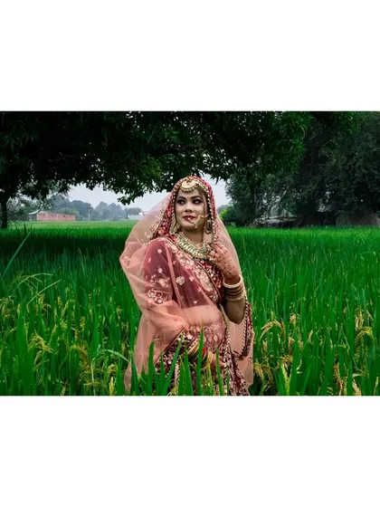 A classic bridal pose showing the full elegance of the wedding lehenga and jewellery. The natural green background makes the traditional red and pink colours of her outfit pop.