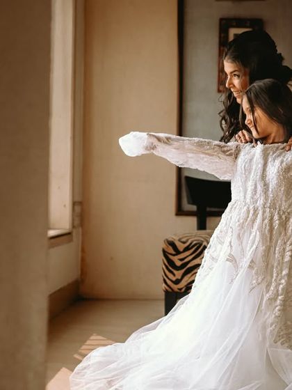 A sister or bridesmaid helps the bride with her dress by the window, a moment of quiet connection.