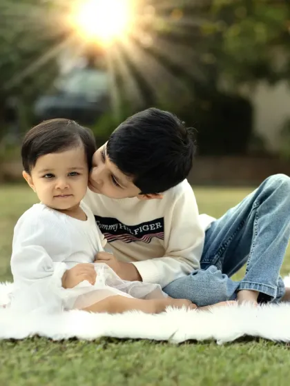 A sweet moment between siblings outdoors, where an older brother gives his baby sister a gentle kiss on the cheek under the warm sun.