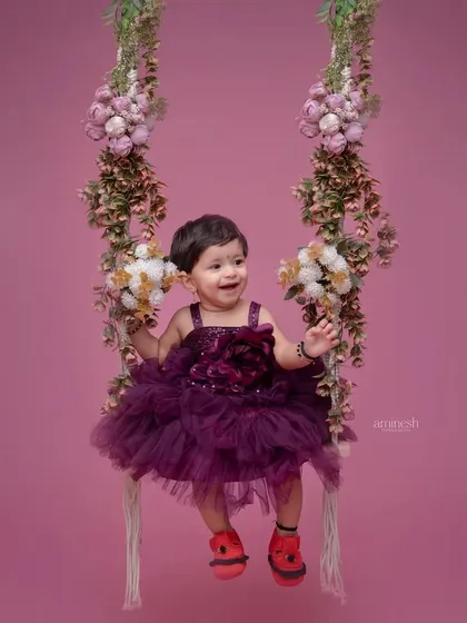 A whimsical portrait of a baby girl in a frilly purple dress on a floral swing. The pink background complements her outfit and creates a sweet, dreamy image.