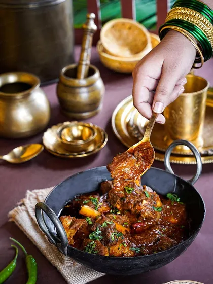 A hand with green bangles takes a spoonful of rich mutton curry from a cast-iron kadai. The rustic, traditional props and moody lighting create a scene straight out of a Maharashtrian home kitchen.