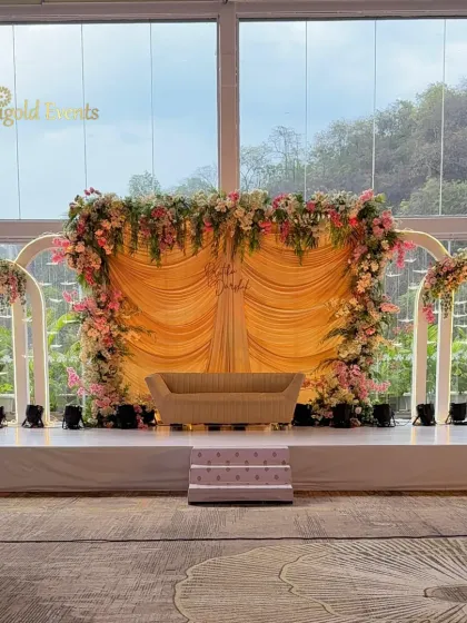 Soft romance against a beautiful natural backdrop. This elegant stage setup features a gracefully draped golden fabric, framed by a lush arrangement of pastel pink and white flowers, perfect for an intimate ceremony or reception.